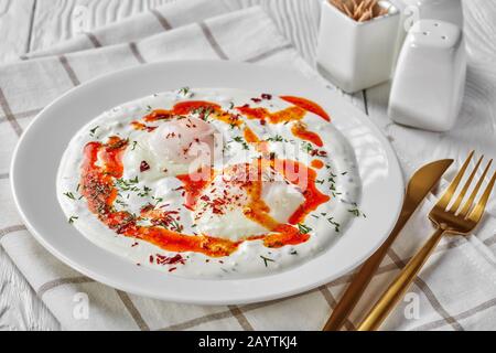 Turkish eggs with greek yogurt and aleppo pepper butter sauce, cilbir, served on white plates with golden cutlery on a white wooden table,  horizontal Stock Photo