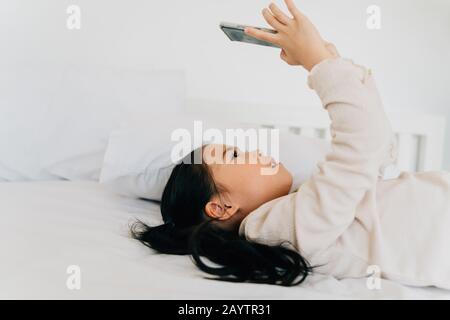 Side view of adorable Asian girl in casual wear playing video game on smartphone lying on bed in house Stock Photo