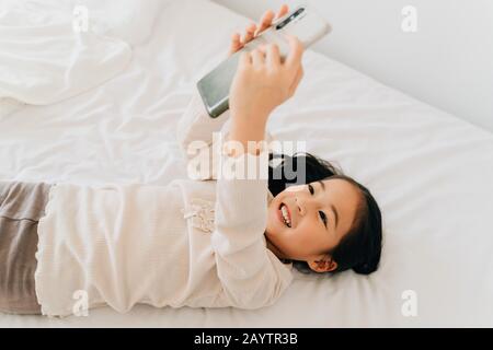 From above of happy smiling Asian girl in casual wear taking selfie on smartphone lying on bed having fun using phone in house Stock Photo