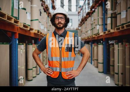 Confident warehouse manager standing in between shelf with hands on waist wearing uniform and hardhat inspecting goods delivery process Stock Photo