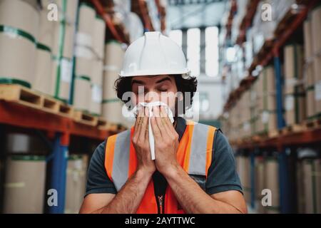 Young warehouse manager coughing and sneezing while feeling sick and covering mouth with handkerchief standing in factory Stock Photo