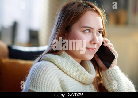 happy modern young woman with red hair in white sweater in the modern house in sunny winter day talking on a cell phone. Stock Photo