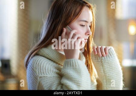 modern teen girl with red hair in white sweater in the modern house in sunny winter day using a smartphone. Stock Photo