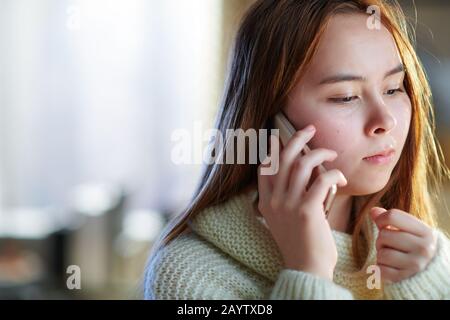 modern young woman with red hair in white sweater in the modern living room in sunny winter day speaking on a mobile phone. Stock Photo