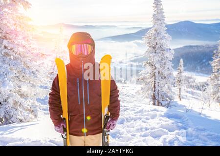 Portrait of skier in protective mask with yellow skis on background of snowy mountains and forest, sunlight Stock Photo