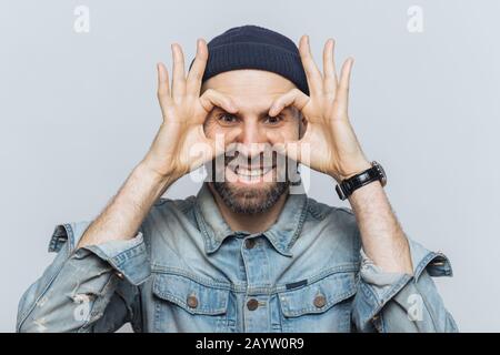 Portrait of hppy bearded male makes eyewear with fingers, has stubble, wears jeans cloting, stands against white background. Funny middle aged man foo Stock Photo