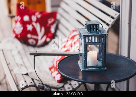 Shot of white candle with burning flame in lamp, stands on round table, bench with cushion in background. Cozy domestic interior. Wonderful decor Stock Photo