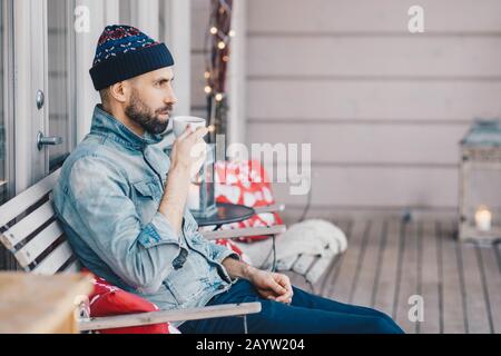Horizontal shot of attractive bearded male wears hat and denim jacket, drinks hot tea, sits at bench on balcony, has thoughtful expression, thinks abo Stock Photo