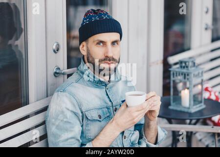 Close up shot of handsome serious bearded male, looks directly into camera with blue eyes and confident expression, drinks hot beverage, enjoys spare Stock Photo