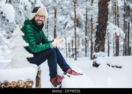 Photo of cheerful male types messages on smart phone looks directly into camera, sits in white winter forest, smiles happily, enjoys communication, wa Stock Photo