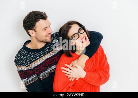 Romantic couple in love embrace each other and have fun together, wear warm knitted sweaters, stand against white background. Cheerful woman and man e Stock Photo
