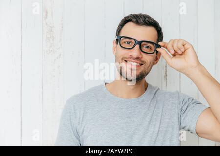 Attractive male model with stubble, looks joyfully through spectacles, has pleasant conversation with someone, wears casual t shirt, poses against whi Stock Photo