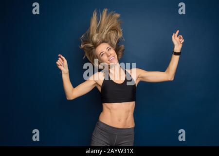 Beautiful smiling fit young woman in sport bra with flowing hair in studio on a blue background. Dance, gym, slim concept. Stock Photo
