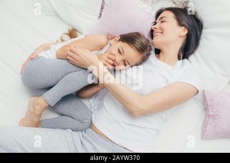 Delighted female child giggles joyfully as plays with her mother in comfortable bed, have positive smiles on faces, wear pyjamas, have good mood after Stock Photo