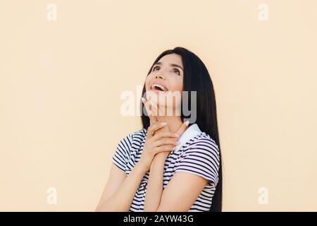 Joyful gorgeous young female model looks happily upwards and keeps hand under chin, notices something pleasant, dressed in casual clothes, wears brace Stock Photo