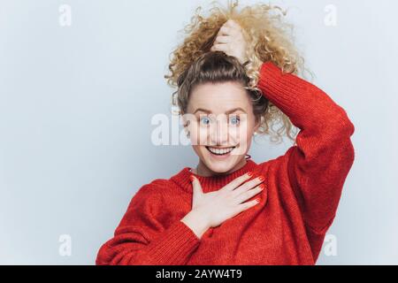 Adorable excited young female with curly hair, keeps hair tied in pony tail, dressed in casual red sweater, happy to recive positive news from interlo Stock Photo