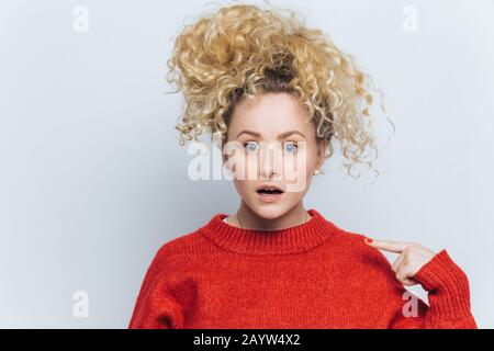 Shocked emotional amazed female stares at camera, indicates at sweater for your advertisment or information, isolated over white background. Surprised Stock Photo