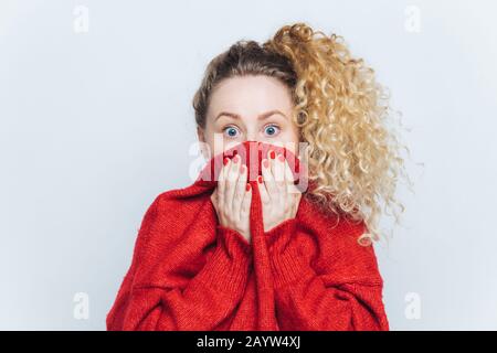 Shocked scared beautiful female stares at camera, being afraid of something awful, covers face with collar, dressed in red loose sweater, isolated ove Stock Photo