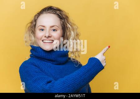 Happy beautiful blonde young woman has pleased expression, wears casual blue sweater, indicates at blank copy space on yellow blank studio wall, adver