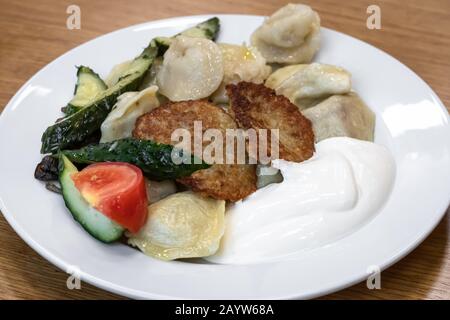 A heap dumplings and fried potato pancakes with sour cream on white plate on wooden background. Traditional Russian dish. Ukrainian cuisine Stock Photo
