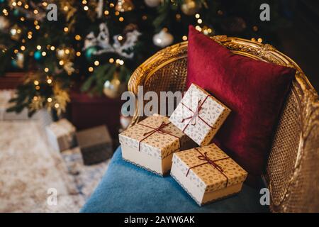 Three wrapped present boxes on armchair near Christmas fir tree. Design or idea for postcard. New Year composition with decorated fir tree and gifts. Stock Photo