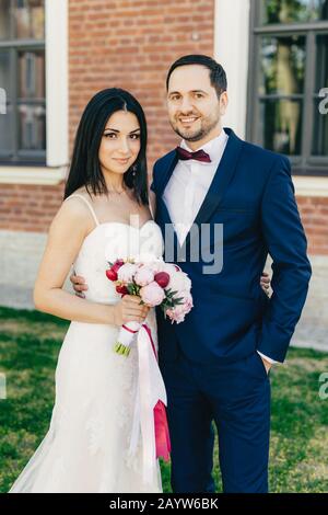 Just married couple embrace each other, have happy expressions, stand close to each other. Beautiful brunette female in white wedding dress, holds bou Stock Photo