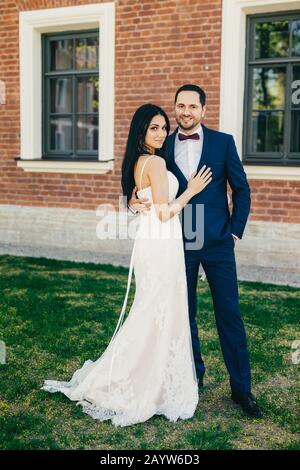 Vertical portrait of attractive bride and handsome bridegroom stand close to each other, hug passionately. Cheerful young wedding couple stand outdoor Stock Photo