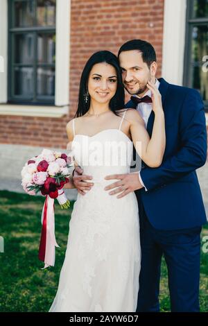 Pleasant looking young female brunette in white wedding dress holds bouquet stands near her handsome bridegroom who wears formal suit, stand outdoors, Stock Photo