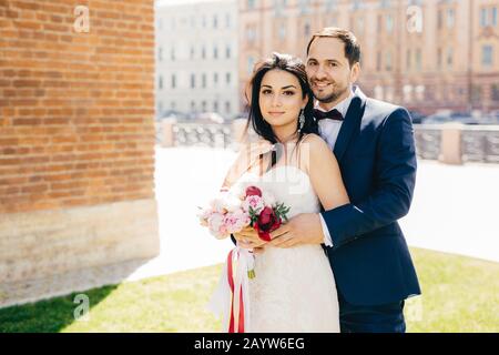 Married couple with happy expressions stand close to each other, embrace passionately, pose outdoors. Beautiful female with appealing appearance, wear Stock Photo