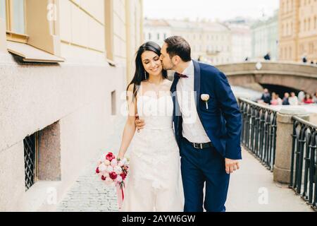 Affectionate handsome bridegroom with beard kisses his future wife. Lovely brunette bride wears nice dress and holds bouquete, has walk with lover on Stock Photo