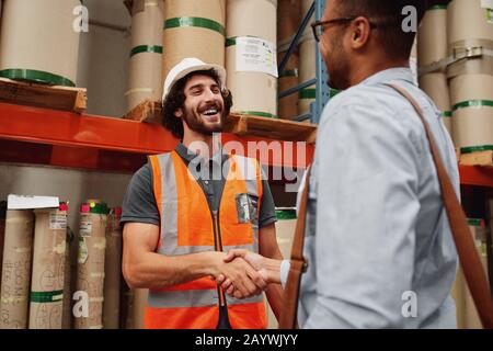 Smiling warehouse manager in orange jacket uniform and helmet greeting business partner and shaking hands Stock Photo