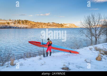 senior male paddler in a drysuit and life jacket is carrying a long unlimited stand up paddleboard on a shore of a lake in Colorado - Horsetooth Reser Stock Photo