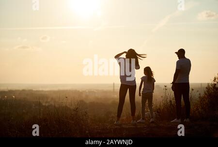 Silhouette of a family standing on the hill with their backs to the camera and looking to the horizon on the sunset, saying goodbye to long and active day Stock Photo