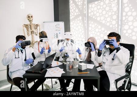 Team of multiethnic pharmacologists wearing protective white coats, gloves and vr goggles, working together on new vaccine or drug in modern Stock Photo