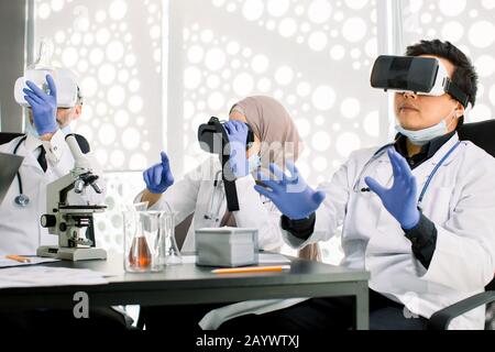 Team of scientists, Asian and Caucasian men, Muslim woman using virtual reality goggles for scientific research in laboratory, gesturing hands Stock Photo