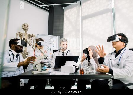 Middle aged male teacher professor has a practical class on microbiology, chemistry with group of multiethnic students, wearing vr goggles while Stock Photo