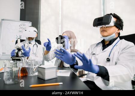 Team of scientists, Asian and Caucasian men, Arabic woman using virtual reality goggles for scientific research in laboratory, gesturing hands Stock Photo