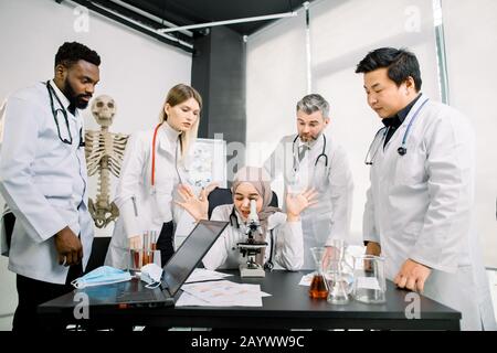 Life scientists researching in laboratory. Female Muslim young scientist microscoping and cheerfully gesturing while her colleagues are standing and