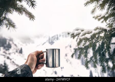Traveler is holding a cup of coffee from in mountains framed with spruce tree at winter time. Mountain camping concept. Stock Photo