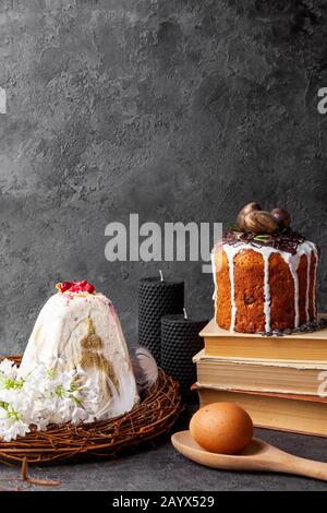 Easter kulich in russian orthodox tradition and baked Easter cake with glaze stands on books, egg on wooden spoon, white flower, black candles, wicker Stock Photo