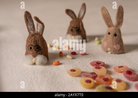 Three brown felt bunnies are sitting between colorful candy eggs on white ground. focus in foreground. Stock Photo