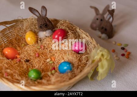 Colored easter eggs and one brown felt bunnies in and two separated other out of the nest of straw with colorful sweet sugar eggs. Felt bunnies to kee Stock Photo