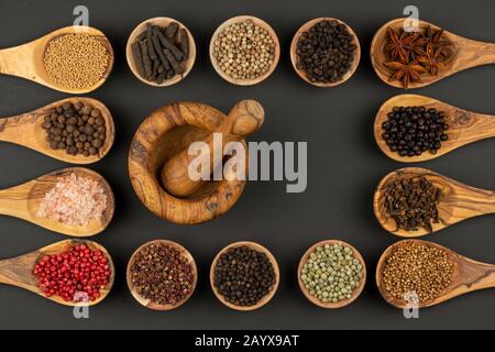 Six small wooden bowls and eight cooking spoons made of olive wood filled with various spices and a mortar of olive wood lie on a black background wit Stock Photo