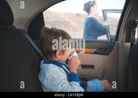 little kid in sweater drinking cup of tea inside car with mother woman calling outside Stock Photo