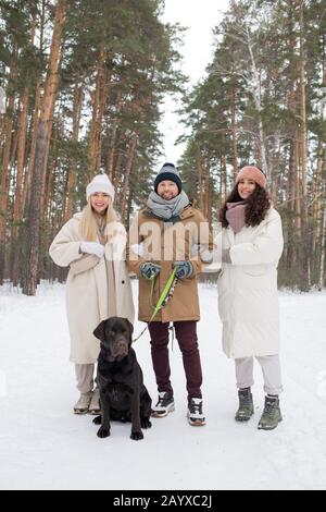 Vertical group shot of young man, two charming women standing together in winter urban forest with dog Stock Photo