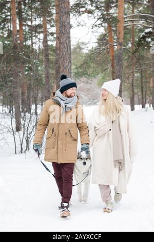 Lovely Caucasian couple chatting while walking their laika dog in urban forest on winter day, vertical shot Stock Photo