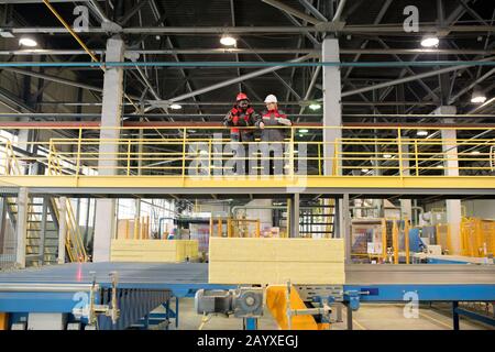 Horizontal long shot of two construction supplies factory workers wearing uniform standing together talking about something Stock Photo