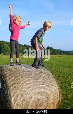 A young children boy and girl (siblings) playing on hay bale summertime. Children summer outdoor activities. Jumping high and far. Concept of summer Stock Photo