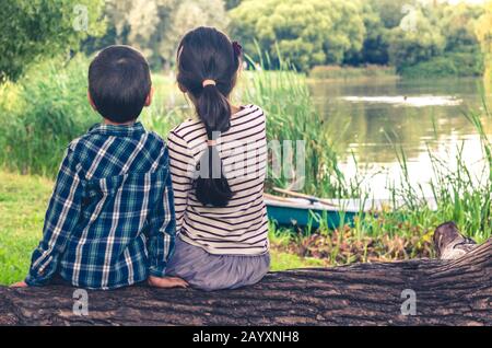 Two children, a boy and a girl, sitting together on a trunk of a tree and watching at lake scenery Stock Photo