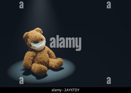 Teddy bear in medical mask, illuminated by spotlight sits in dark room. Concept of Infectious diseases such as Coronavirus Stock Photo